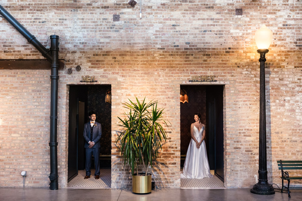 A bride and groom stand in separate restroom doorways against a brick wall, facing the camera, capturing a quirky moment from their Wildman BT wedding.
