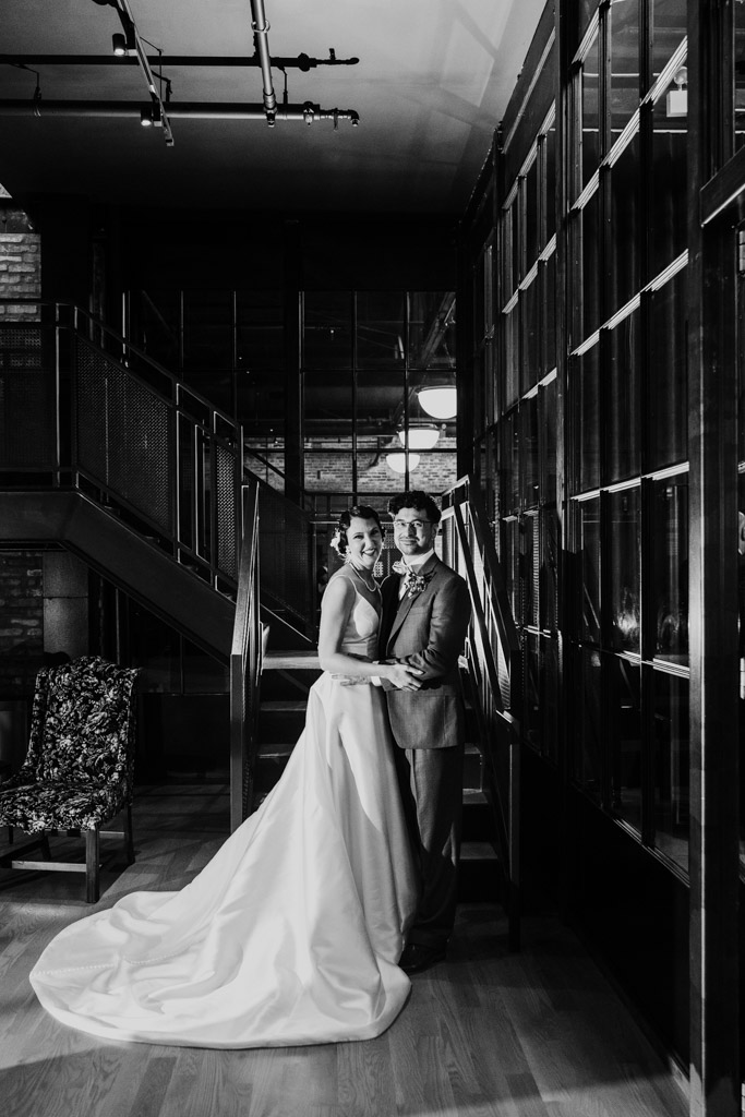 A bride and groom stand together indoors near a staircase, dressed in wedding attire, in black and white at a Wildman BT wedding.