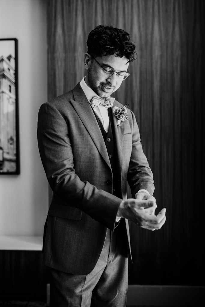 A man in a suit adjusts his cufflink, standing indoors in front of a textured wall—perhaps preparing for a Wildman BT wedding.