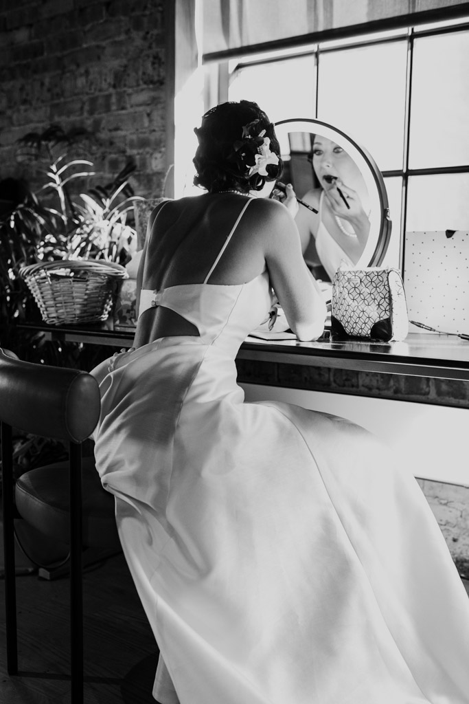 A bride in a white dress, preparing for her Wildman BT wedding, applies lipstick at a vanity table, seen from behind in black and white.