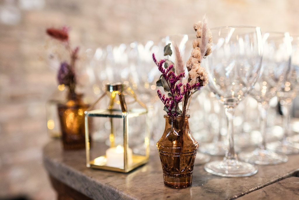 Small brown vases with dried flowers and a lantern add rustic charm beside rows of empty wine glasses on the Wildman BT wedding countertop.