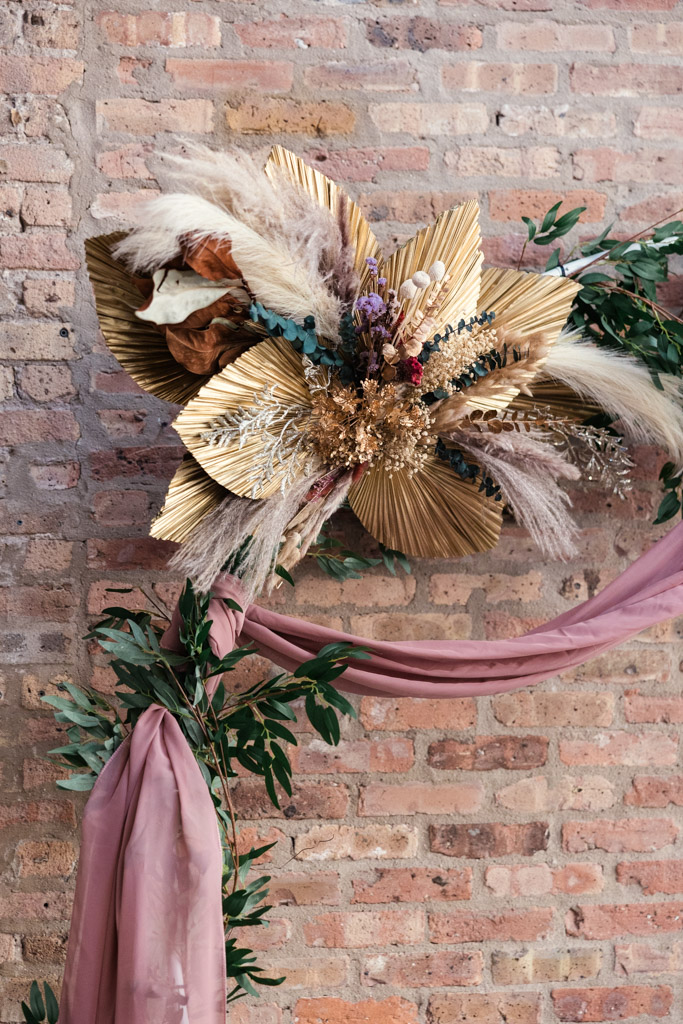 Dried floral arrangement with gold leaves and pampas grass on a brick wall, draped with mauve fabric and greenery—perfect for a Wildman BT wedding backdrop.