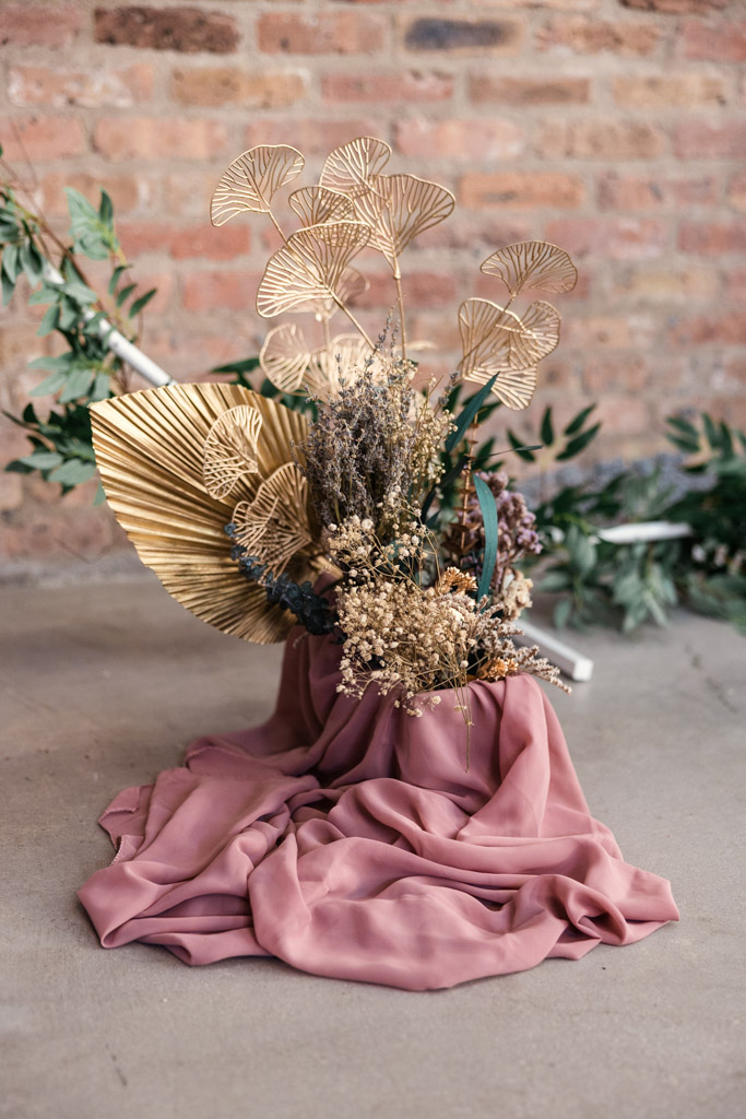 Dried floral arrangement with gold leaves on pink fabric, set against a brick wall and concrete floor—perfect for a Wildman BT wedding.