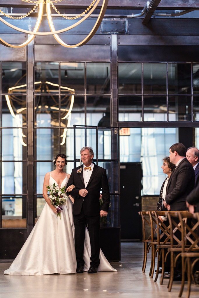 At a Wildman BT wedding, a bride in a white dress walks down the aisle with an older man, both smiling, at an indoor ceremony.