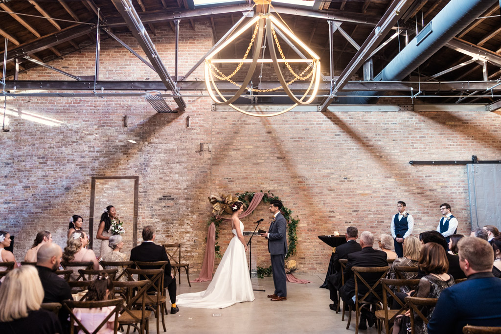 A bride and groom exchange vows at an indoor Wildman BT wedding ceremony, with guests seated on either side.
