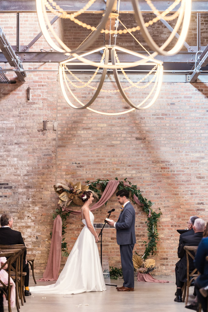 A bride and groom stand together at a Wildman BT wedding ceremony in front of a floral arch and exposed brick wall.