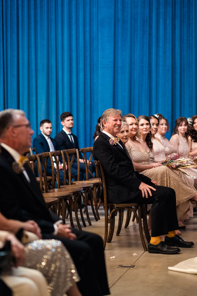 Guests in formal attire sit on wooden chairs, smiling, in front of a blue curtain at the Wildman BT wedding indoor event.