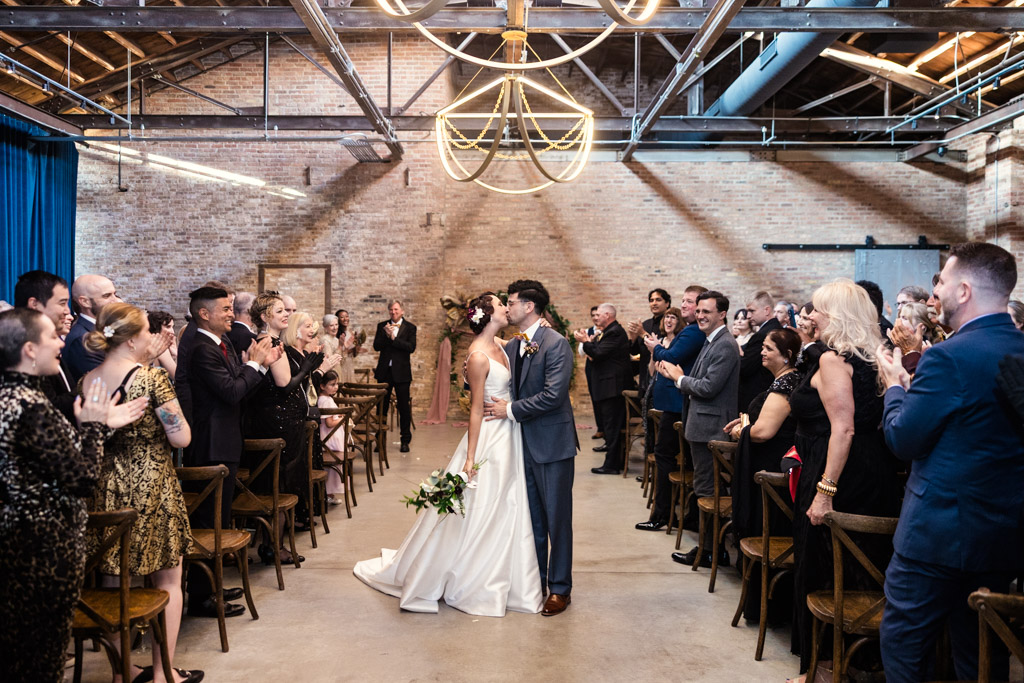 A bride and groom share a kiss at the end of the aisle as wedding guests applaud in an indoor venue during their beautiful Wildman BT wedding.