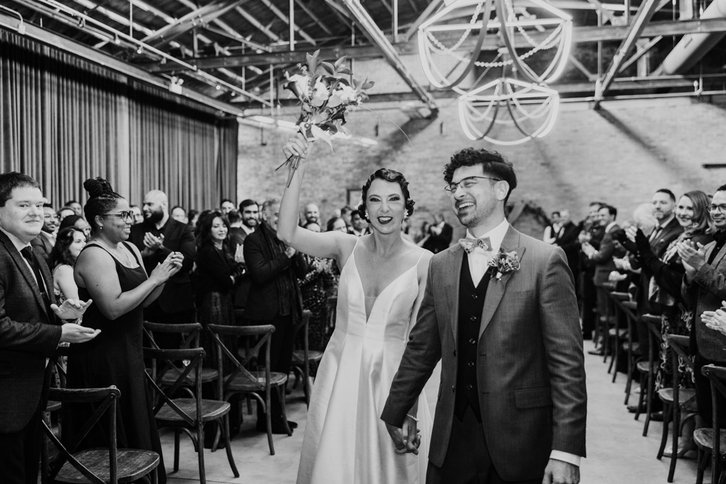 Bride and groom walk down the aisle smiling at their Wildman BT wedding, guests applaud; bride holds bouquet up in celebration.