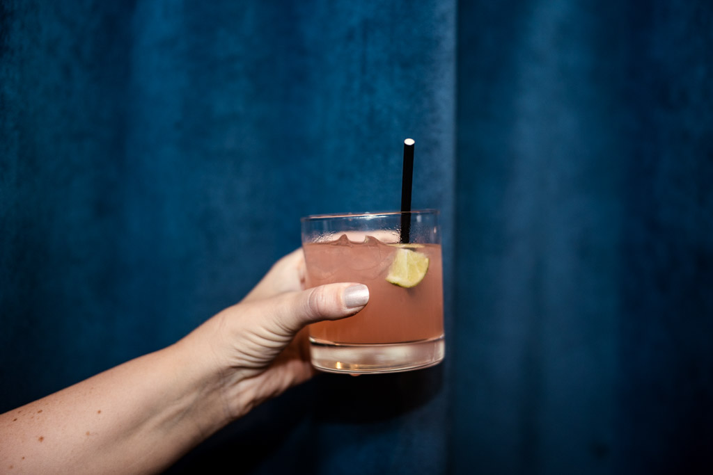 A hand holds a pink cocktail with ice, a lime wedge, and a straw against a dark blue background, perfect for raising a toast at the Wildman BT wedding.