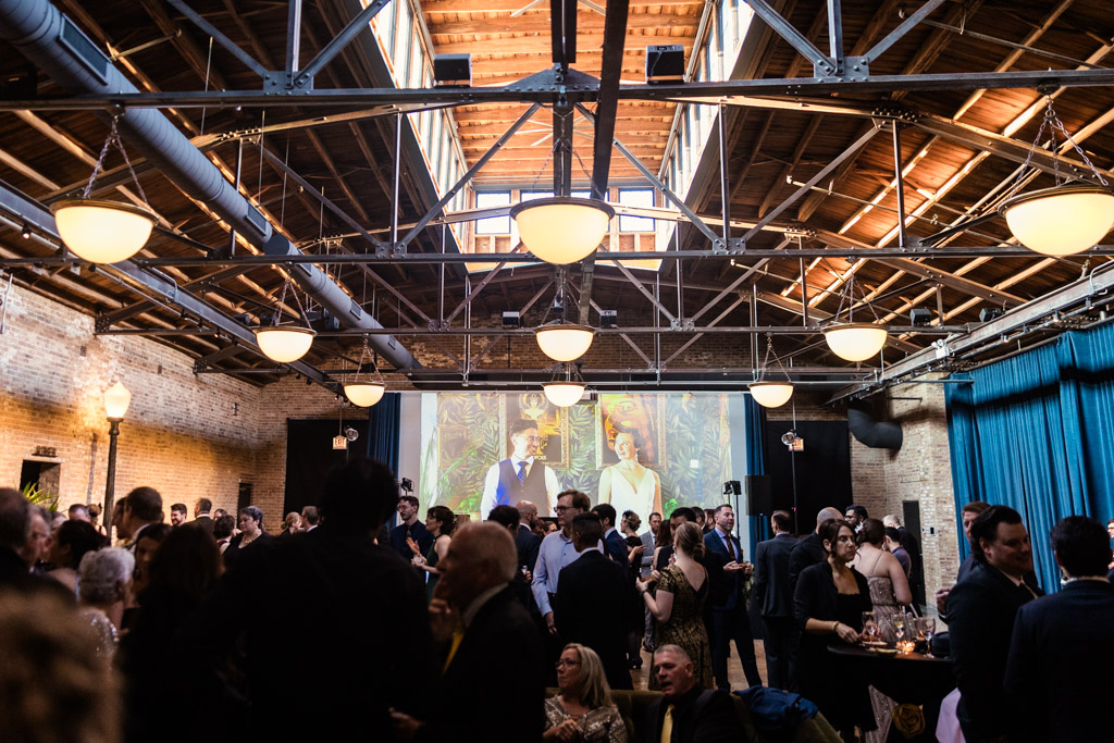 A large group of people gather at the Wildman BT wedding, an indoor event with high ceilings and a projected screen in the background.