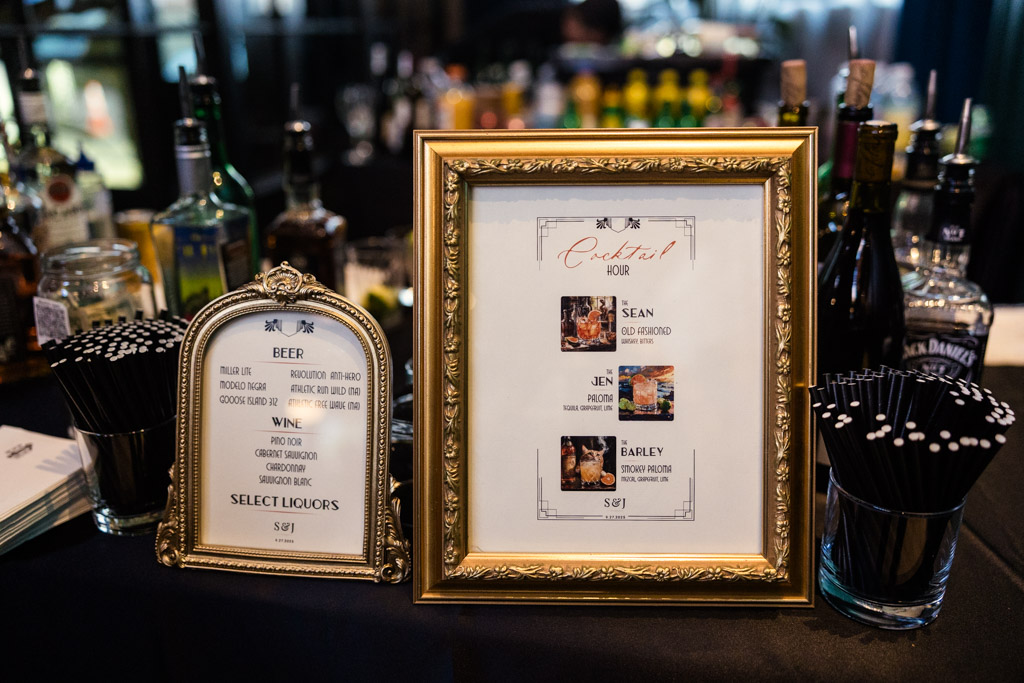 Framed bar menus for beer, wine, and cocktails displayed on a counter with various bottles and black straws at a Wildman BT wedding.
