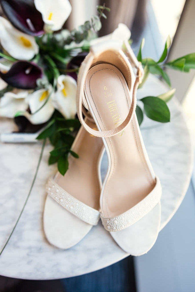 A pair of elegant white sandals on a marble table with a bouquet of white and dark purple flowers—perfect for a Wildman BT wedding.