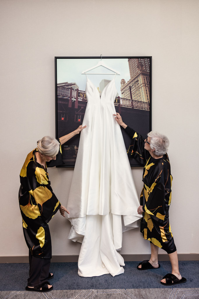 Two women admire a stunning white wedding dress, part of the Wildman BT wedding collection, as it hangs gracefully on a framed photo of a cityscape.