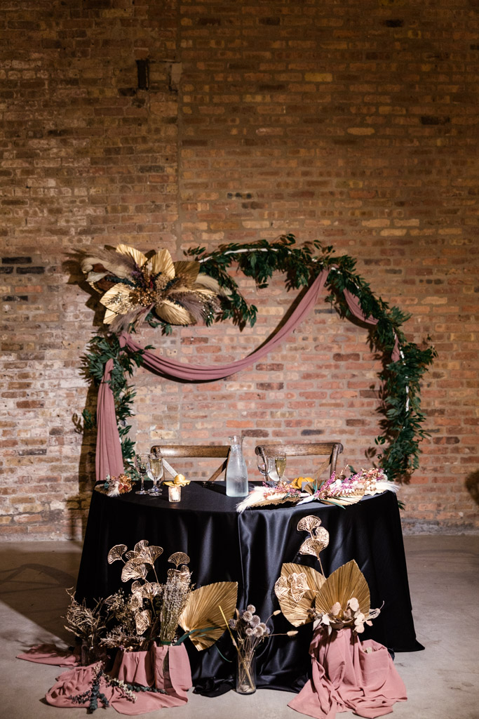 A Wildman BT wedding sweetheart table with black cloth, gold accents, and greenery sits in front of a brick wall adorned with elegant decor.