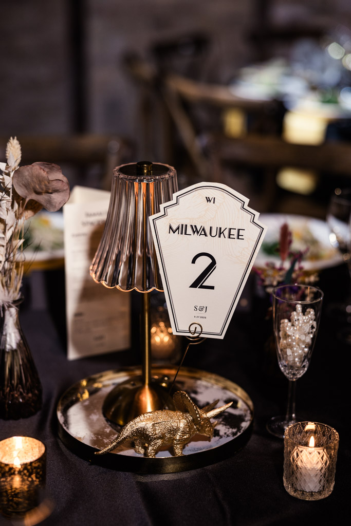 Table centerpiece for a Wildman BT wedding featuring a Milwaukee table number sign, lamp, candles, dried flowers, and a gold dinosaur figurine.