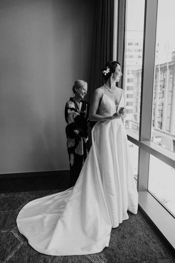 A bride in a gown stands by a window with an older woman behind her, both looking outside before the Wildman BT wedding.