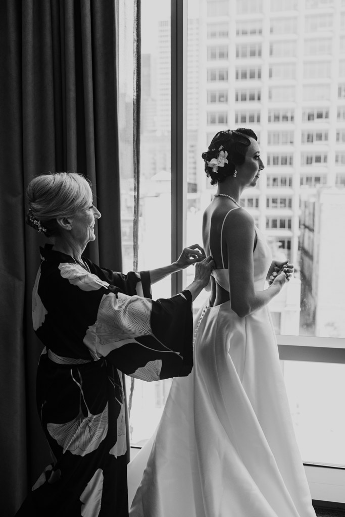 A woman fastens a bride’s dress by a window overlooking city buildings, capturing a quiet moment before the Wildman BT wedding.