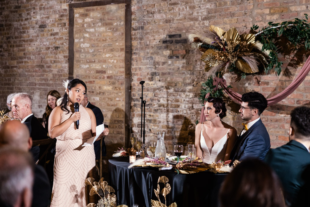 A woman gives a heartfelt speech at a Wildman BT wedding reception as the bride and groom listen lovingly at their table.