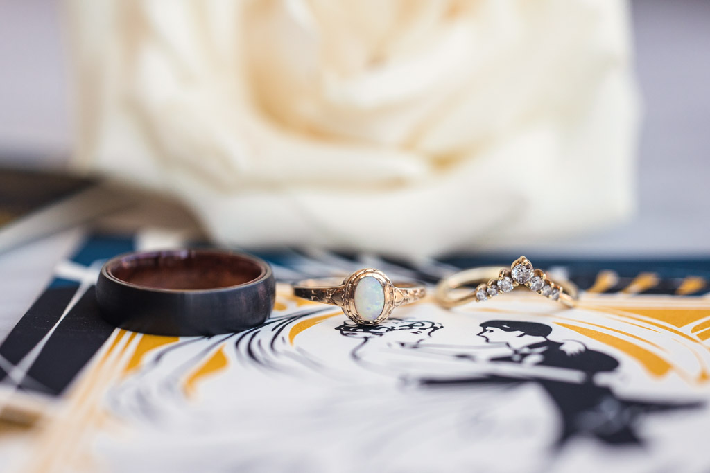 Three rings in front of a white rose, with a Wildman BT wedding invitation featuring a dancing couple in the background.