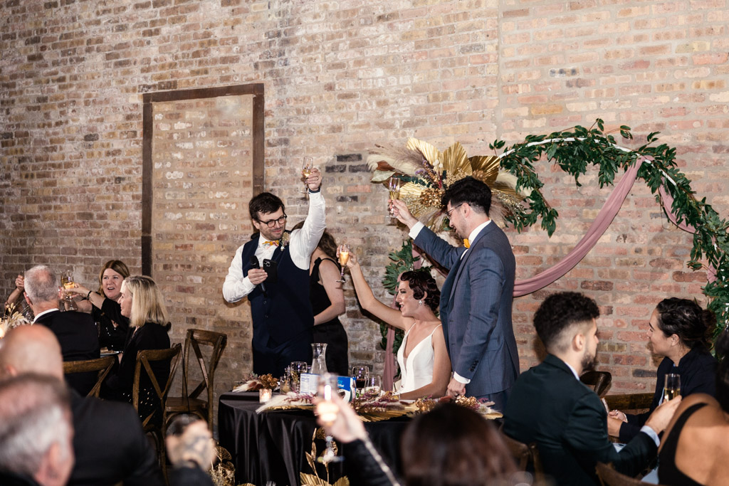 Guests and newlyweds raise glasses for a toast at a Wildman BT wedding, set in a rustic, brick-walled venue.