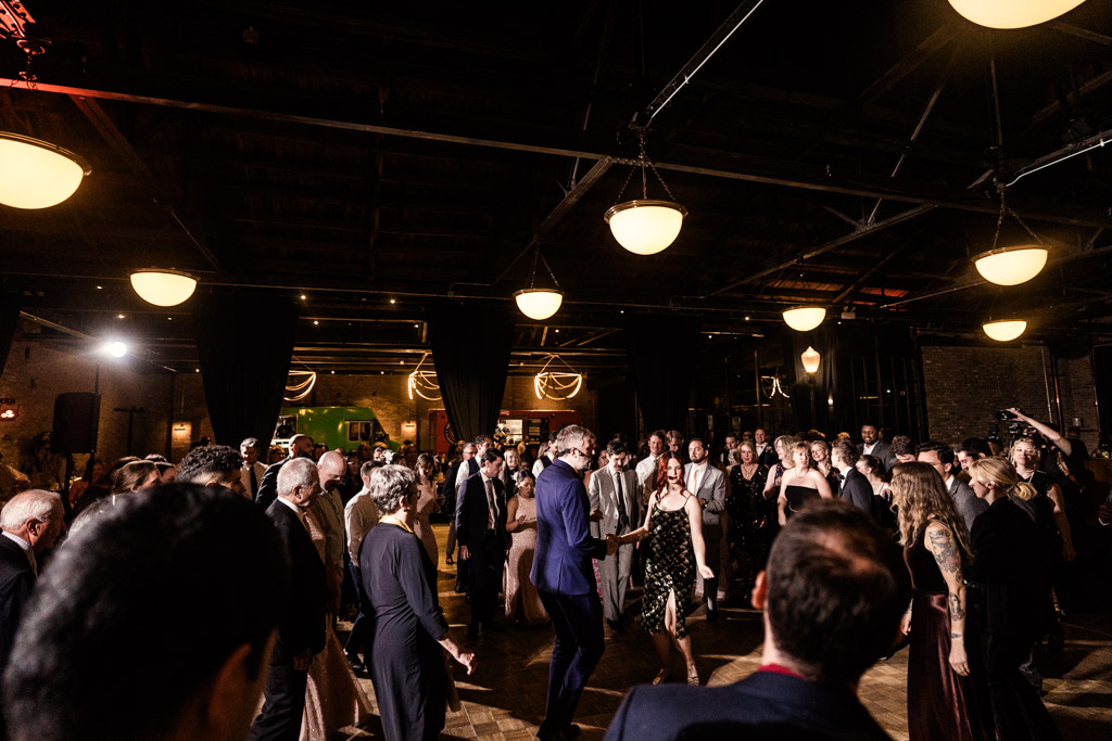 A large group of people dance together at a lively indoor Wildman BT wedding under warm hanging lights.