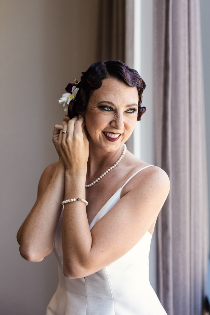 Smiling bride in a white dress adjusts her earring by a window at her Wildman BT wedding, wearing a pearl necklace and bracelet.