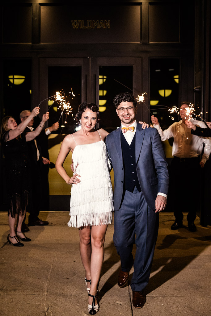 A smiling couple walks outside at night during a Wildman BT wedding as people behind them hold sparklers and celebrate.
