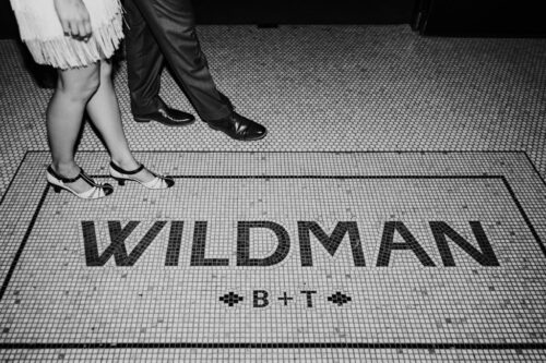 A man and a woman stand on a tiled floor with "WILDMAN B+T" written on it, capturing a special moment from the Wildman BT wedding.