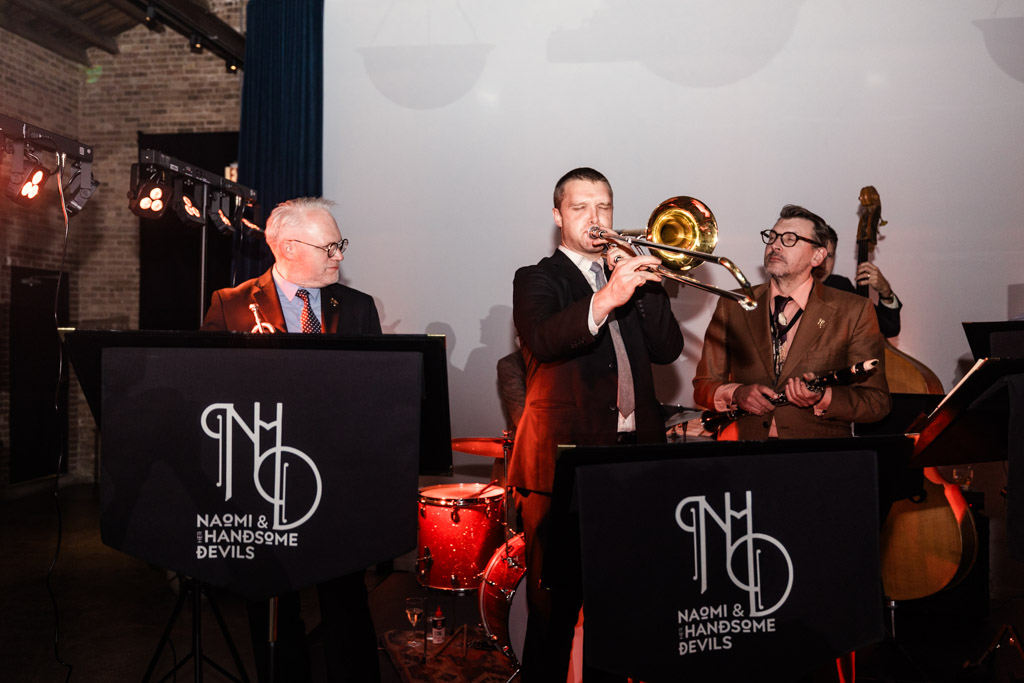 Three musicians perform on stage at the Wildman BT wedding; one plays trombone, while the others play saxophone and stand behind music stands.