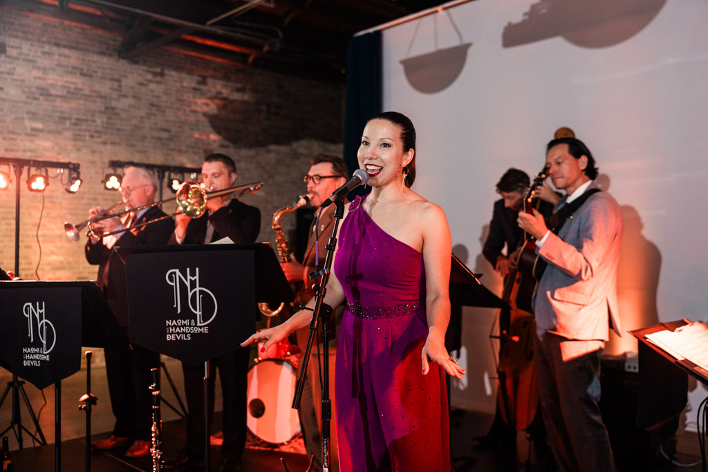 At a Wildman BT wedding, a woman in a purple dress sings with a jazz band playing instruments on a dimly lit stage.