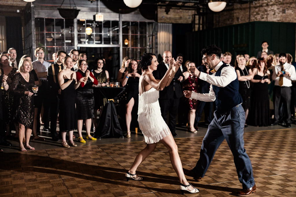 A couple dances energetically on a wooden floor at a Wildman BT wedding, while guests watch and smile at the lively event.