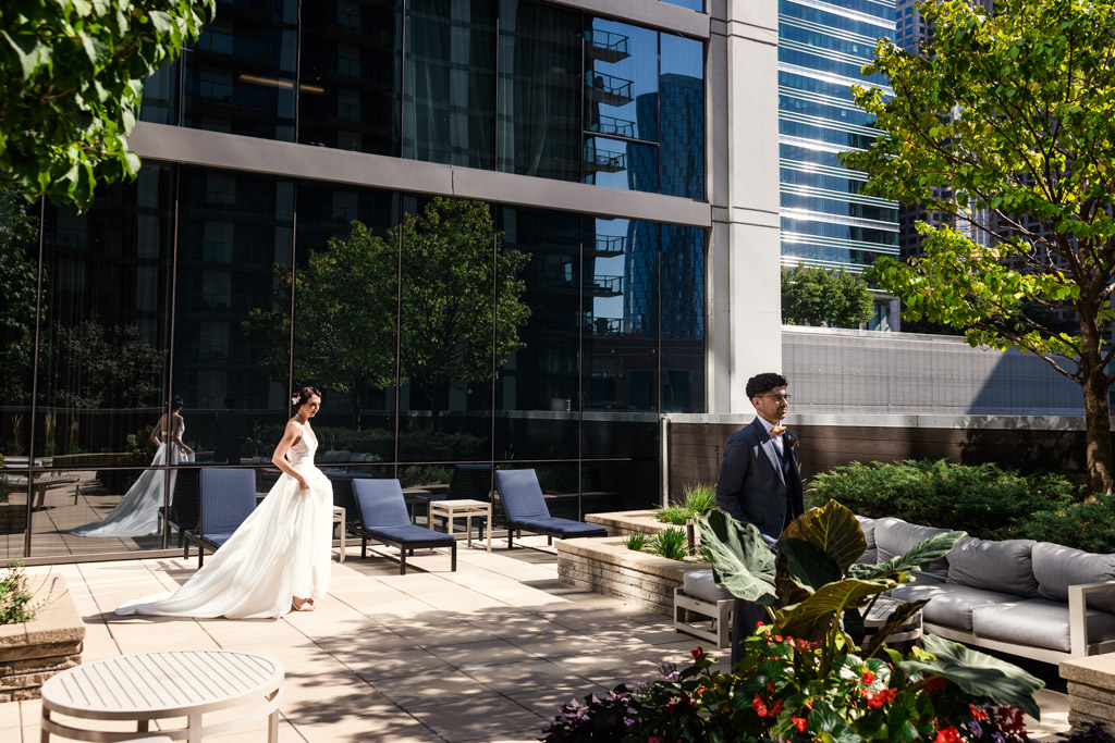 A bride in a white dress approaches her groom outdoors on a modern city terrace with trees and lounge chairs, capturing the magic of their Wildman BT wedding.