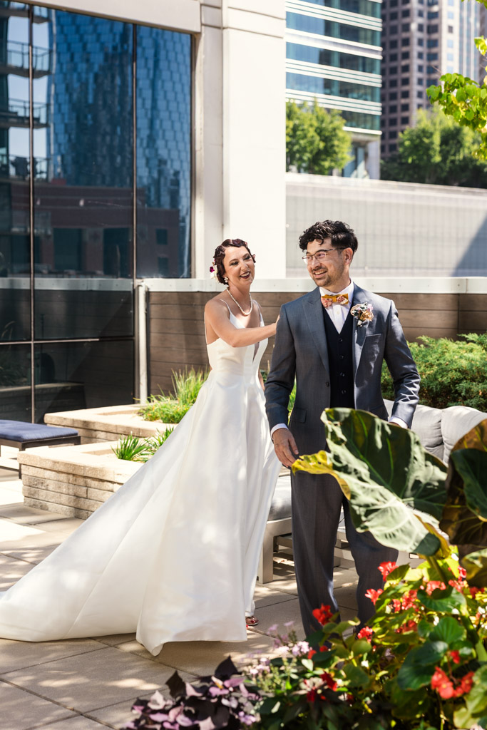 A bride in a white dress taps a smiling groom’s shoulder during their Wildman BT wedding, set outside in a modern urban setting.