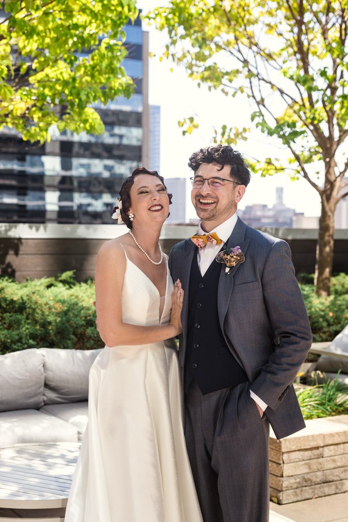 A bride and groom smile outdoors in elegant wedding attire, greenery and city buildings behind them, capturing the joyful spirit of a Wildman BT wedding.