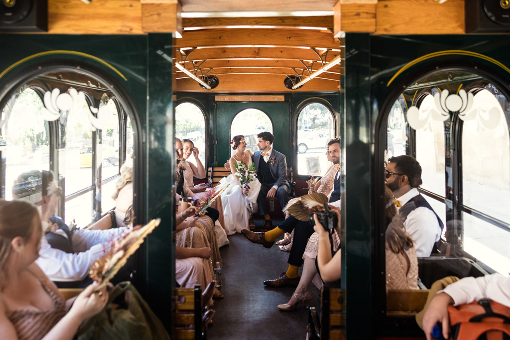 A bride and groom sit together on a trolley at their Wildman BT wedding, surrounded by their wedding party dressed in formal attire.