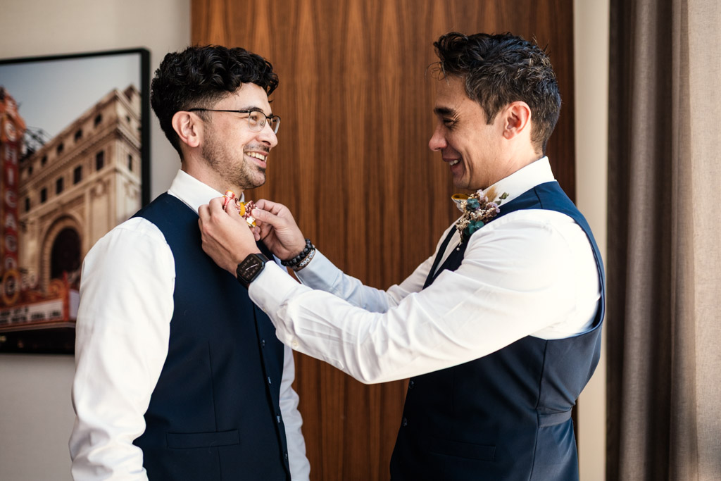 Two men in vests smile warmly as one helps the other adjust his bowtie in a bright room, capturing a joyful moment at a Wildman BT wedding.