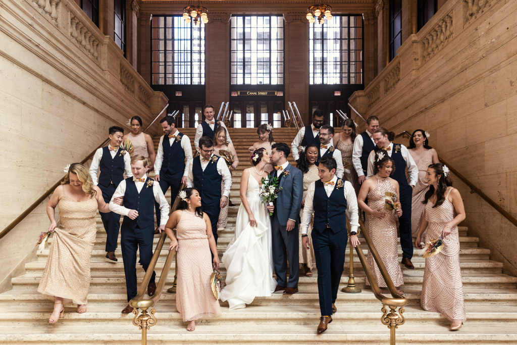 A bride and groom share a kiss while surrounded by their Wildman BT wedding party on a grand staircase indoors.