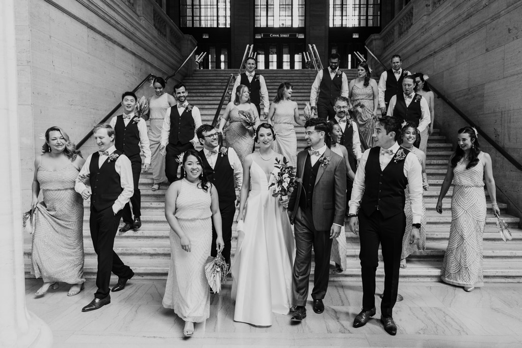 A Wildman BT wedding party walks down marble stairs, with the bride and groom in the center.
