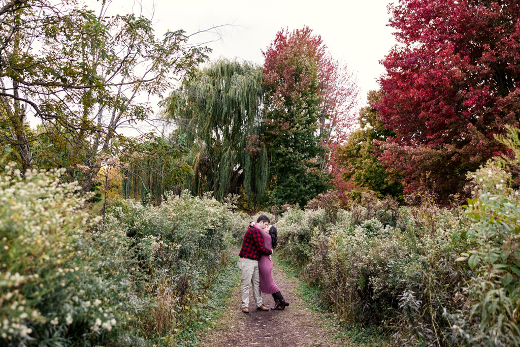 Engaged couple embraces on a leafy path surrounded by tall, colorful autumn trees and foliage in Winnemac Park in Chicago