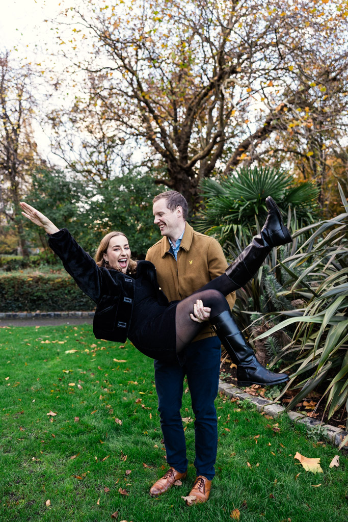 Man lifts his happy fiance woman in Merrion Square, surrounded by green grass and autumn trees during Dublin engagement session