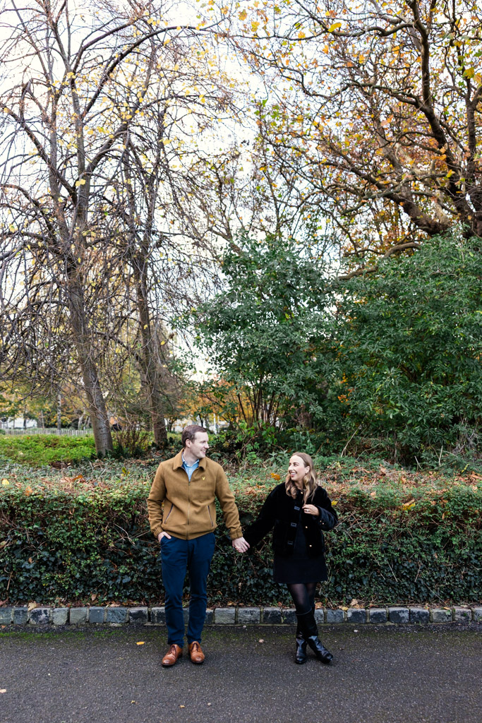 Engaged couple holding hands and smiling while walking outdoors on a path in Merrion Square, surrounded by autumn trees and greenery, during their Dublin engagement session