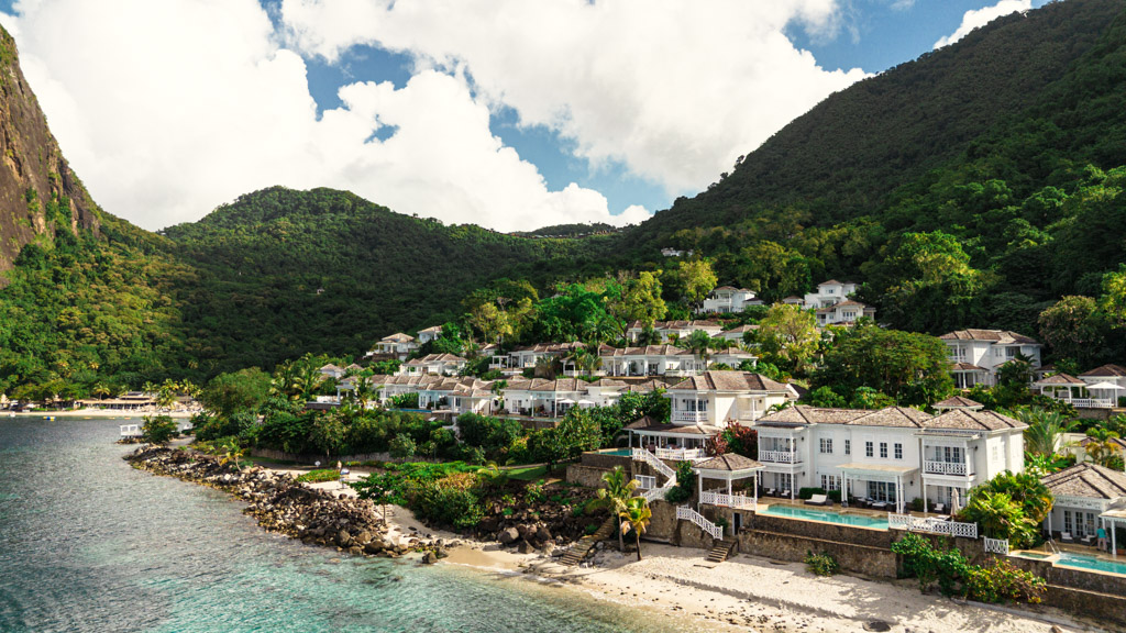 White villas at Viceroy Sugar Beach nestle on a lush green hillside by the beach and clear blue water, with mountains in the background