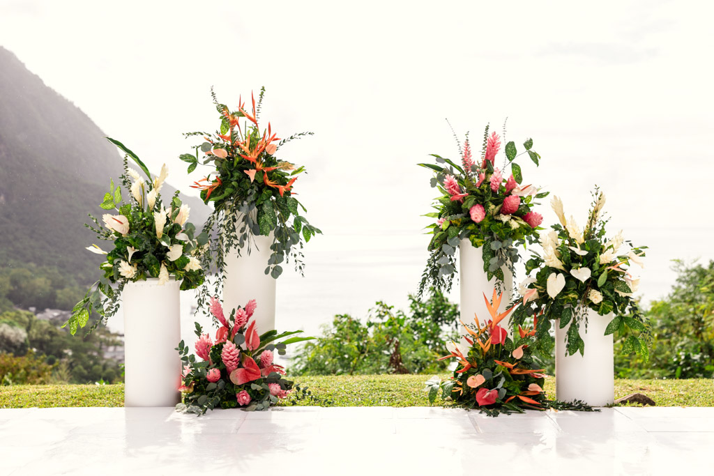 Four tall white vases filled with tropical flowers and greenery, beautifully arranged outdoors near a mountain and ocean for wedding ceremony at Viceroy Sugar Beach