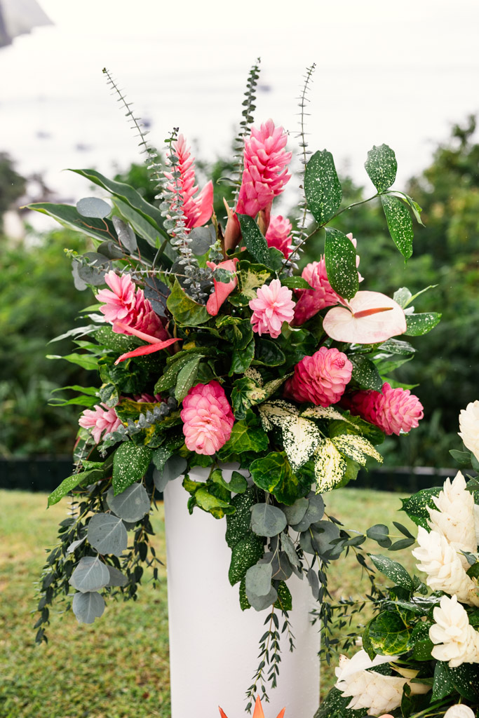 A tall white vase holds a vibrant arrangement of pink tropical flowers and lush green leaves outdoors for Viceroy Sugar Beach wedding ceremony