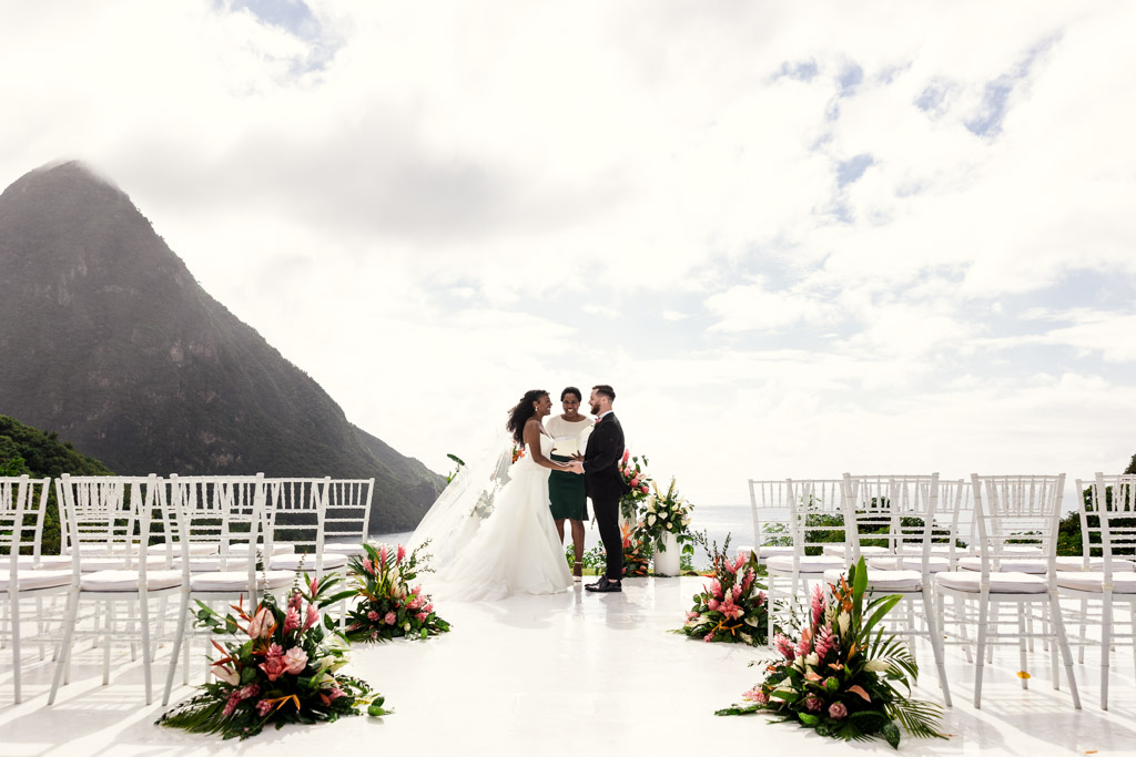 Bride and groom stand with officiant outdoors at Viceroy Sugar Beach, mountains in the background, surrounded by white chairs and flowers