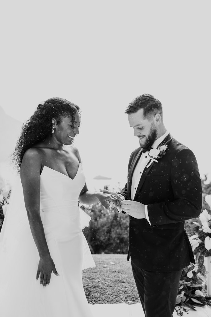 Black and white photo of bride and groom smiling as the groom places a ring on the bride's finger during their outdoor wedding ceremony at Viceroy Sugar Beach