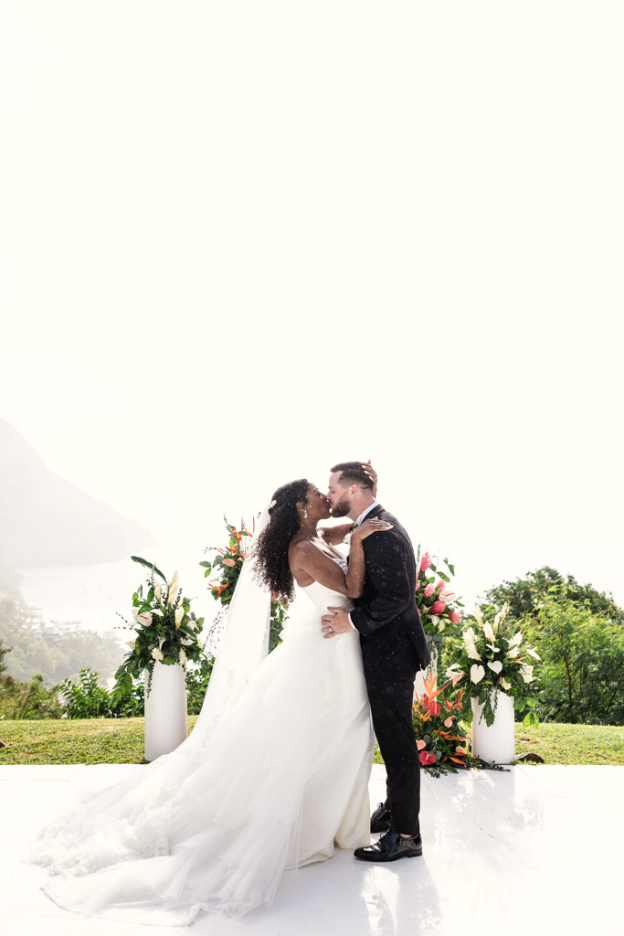 A bride and groom kiss outdoors at Viceroy Sugar Beach, surrounded by flowers, with a scenic, misty background