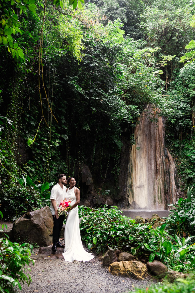 A couple in wedding attire stands together at Diamond Falls, surrounded by lush green jungle foliage