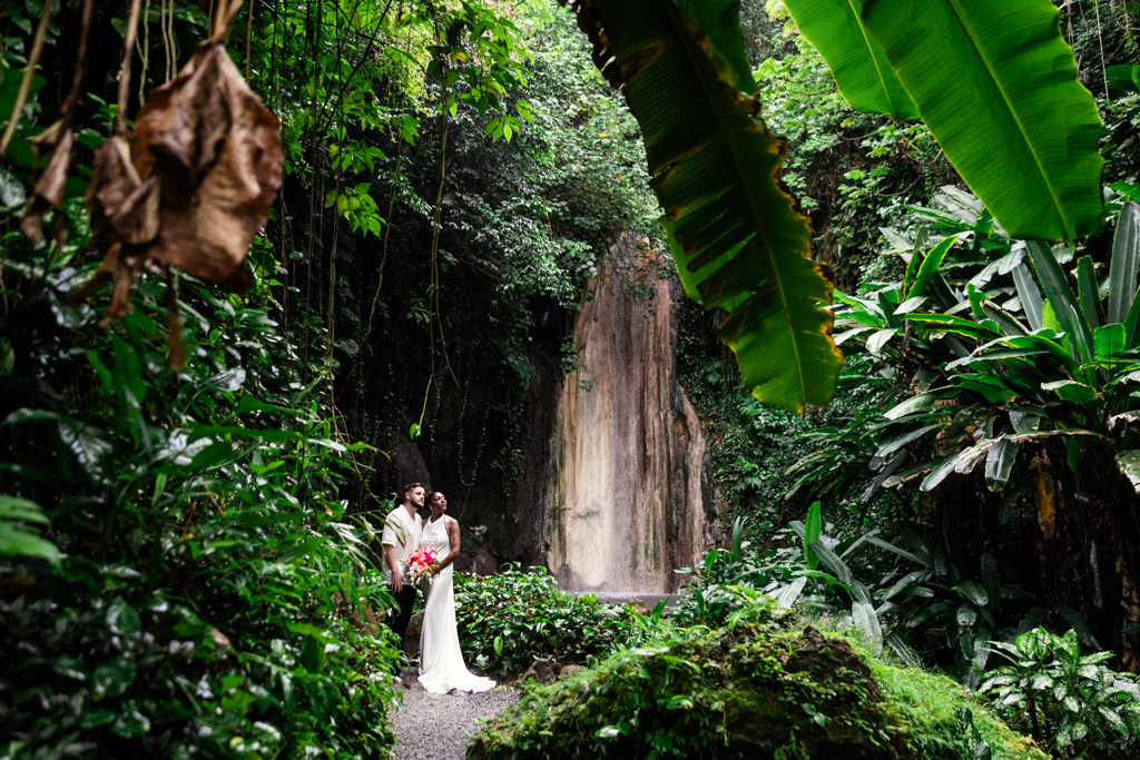 Bride and groom in wedding attire stans by a waterfall at Diamond Falls Botanical Gardens, surrounded by lush green tropical plants